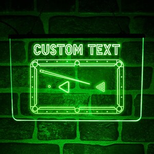 Può includere: Un'insegna al neon a LED rettangolare che brilla di un verde brillante. L'insegna presenta il testo CUSTOM TEXT nella parte superiore, con un grafico di un tavolo da biliardo, una stecca, un triangolo e palle da biliardo centrati.