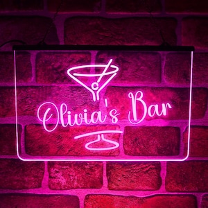 May include: A pink neon sign displaying the words "Olivia's Bar" in a cursive font, with a martini glass icon. The sign is rectangular and mounted on a brick wall.