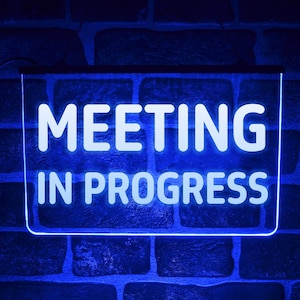 Może przedstawiać: Prostokątny, podświetlany znak z białym napisem "MEETING IN PROGRESS", obramowanym na niebiesko. Znak jest zamontowany na ciemnoniebieskiej ceglanej ścianie, tworząc kontrast.
