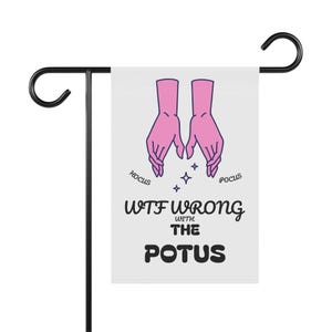 Könnte beinhalten: Eine weiße Gartenflagge auf einem schwarzen Metallständer. Die Flagge zeigt rosa Hände mit den Worten "Hocus Pocus" und "WTF Wrong with the POTUS" in schwarzem Text. Vier kleine Sterne befinden sich zwischen den Händen.