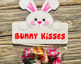 May include: A white bunny peeking over a sign that says "Bunny Kisses". A clear plastic bag filled with colorful foil-wrapped chocolate kisses.