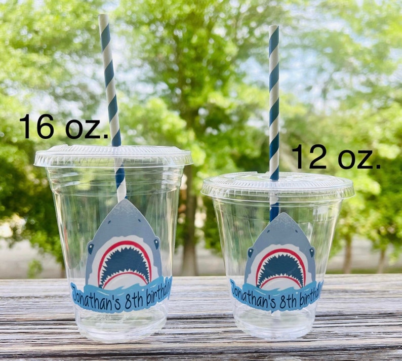 May include: Two clear plastic cups with blue and white striped straws. The cups have a shark graphic and the text "Jonathan's 8th birthday". The larger cup is 16 fl oz and the smaller cup is 12 fl oz.