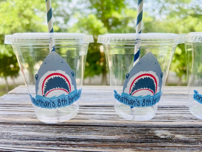May include: Clear plastic cups with blue and white striped straws. Each cup has a shark sticker with the text "Jonathan's 8th birthday" on it.