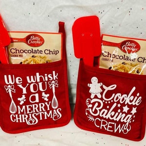 May include: Two red fabric oven mitts with white text and a small red spatula and a box of Betty Crocker chocolate chip cookie mix in each. The left mitt says "We whisk you a merry Christmas" and the right mitt says "Cookie Baking Crew".