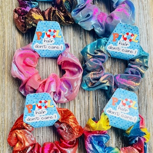 May include: Six iridescent hair scrunchies with a holographic finish. Each scrunchie has a tag that reads "Pool hair don't care!" The scrunchies are in the colors purple, brown, blue, pink, red, and rainbow.
