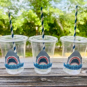May include: Three clear plastic cups with blue and white striped straws. Each cup has a shark sticker with the text "Jonathan's 8th birthday".