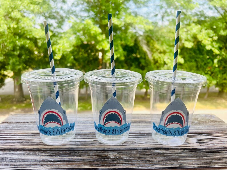 May include: Three clear plastic cups with blue and white striped straws. Each cup has a shark sticker with the text "Jonathan's 8th birthday".