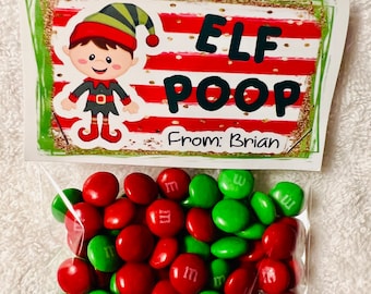Puede incluir: Una pequeña bolsa de plástico transparente llena de caramelos M&M's rojos y verdes. Una tarjeta blanca con un borde verde y rayas rojas y blancas está unida a la bolsa. La tarjeta tiene un duende de dibujos animados y las palabras "ELF POOP" en letras negras. La tarjeta también dice "De: Brian" en letras doradas.