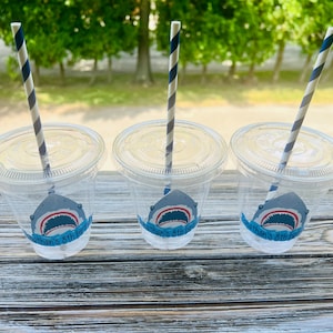 May include: Three clear plastic cups with blue and white striped straws. Each cup has a shark-themed sticker with the text "Nathan's 8th Birthday".