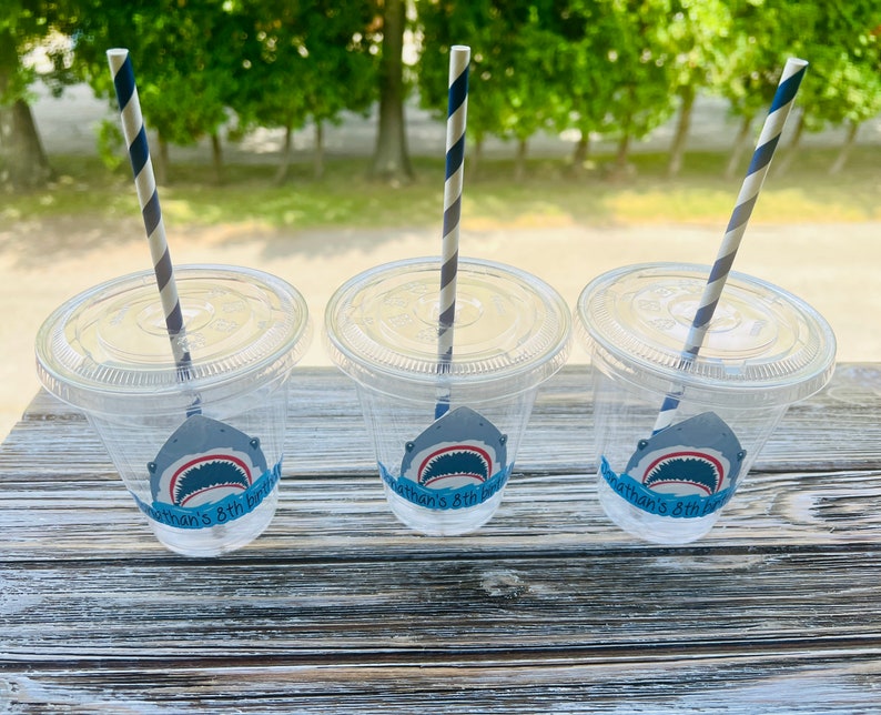 May include: Three clear plastic cups with blue and white striped straws. Each cup has a shark-themed sticker with the text "Nathan's 8th Birthday".