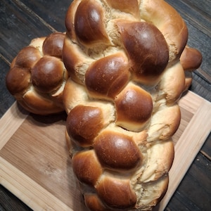 May include: Two golden-brown braided loaves of challah bread on a wooden cutting board. The bread has a glossy, baked crust and a soft, fluffy interior. The loaves are arranged side-by-side, showcasing the intricate braiding.