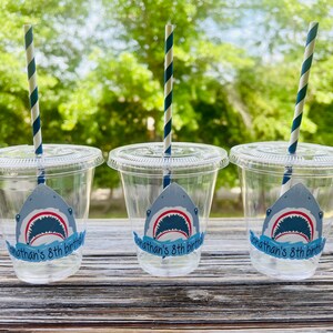 May include: Three clear plastic cups with blue and white striped straws. Each cup has a shark sticker with the text "Jonathan's 8th birthday".