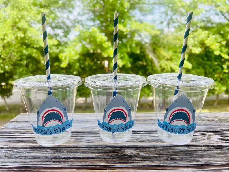 May include: Three clear plastic cups with blue and white striped straws. Each cup has a shark sticker with the text "Jonathan's 8th birthday".