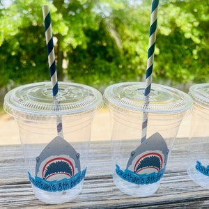 May include: Three clear plastic cups with blue and white striped straws. Each cup has a shark sticker with the text "Jonathan's 8th birthday" on it.