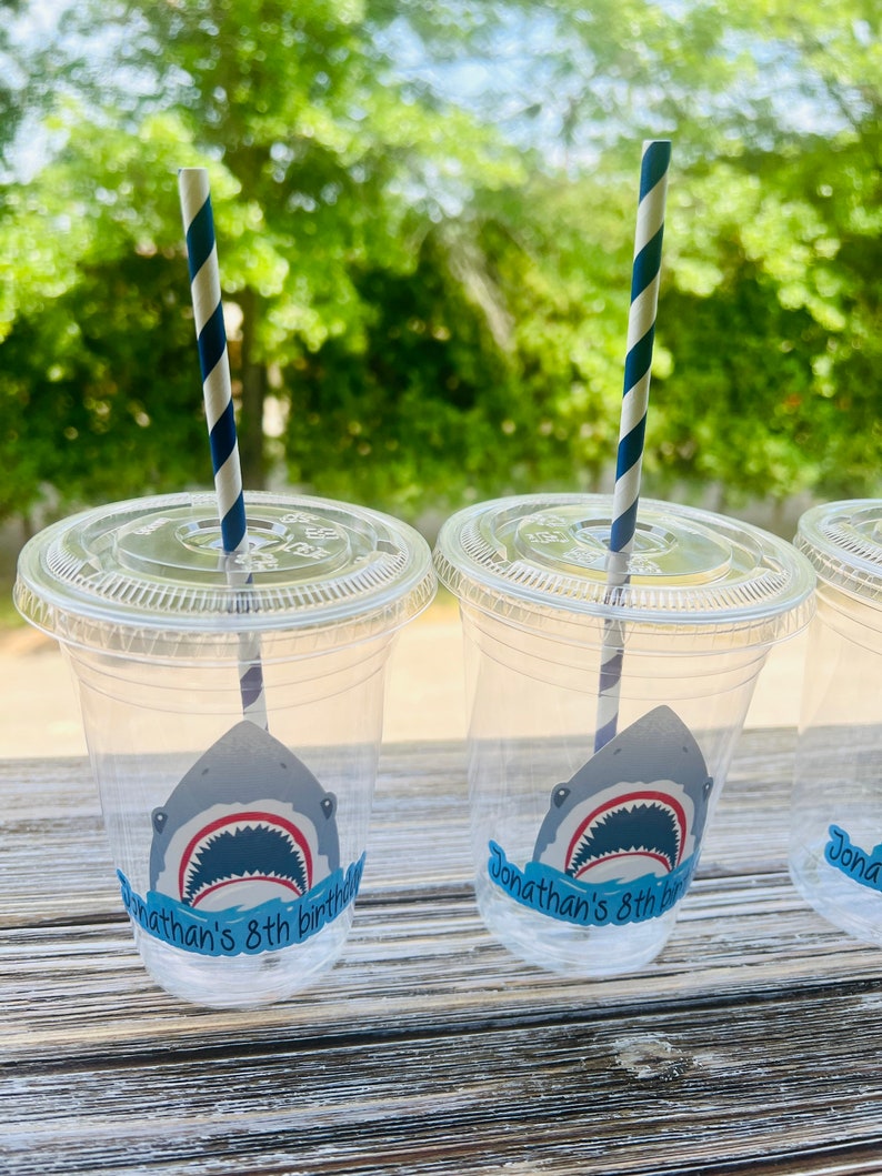 May include: Three clear plastic cups with blue and white striped straws. Each cup has a shark sticker with the text "Jonathan's 8th birthday" on it.