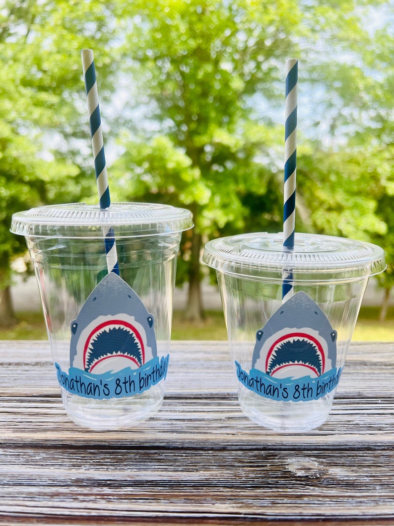May include: Two clear plastic cups with blue and white striped straws. Each cup has a shark sticker with an open mouth and the text "Jonathan's 8th birthday".