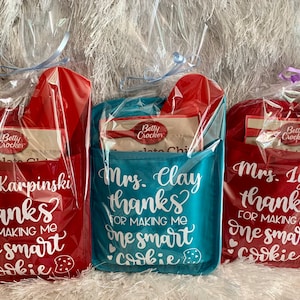 May include: Three red and blue fabric gift bags with the text "thanks for making me one smart cookie" and the teacher's name. Each bag includes a box of Betty Crocker chocolate chip cookie mix and a red kitchen utensil.
