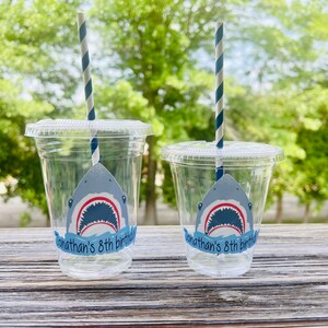 May include: Two clear plastic cups with blue and white striped straws. Each cup has a shark sticker with an open mouth and the text "Jonathan's 8th birthday" on a blue banner.