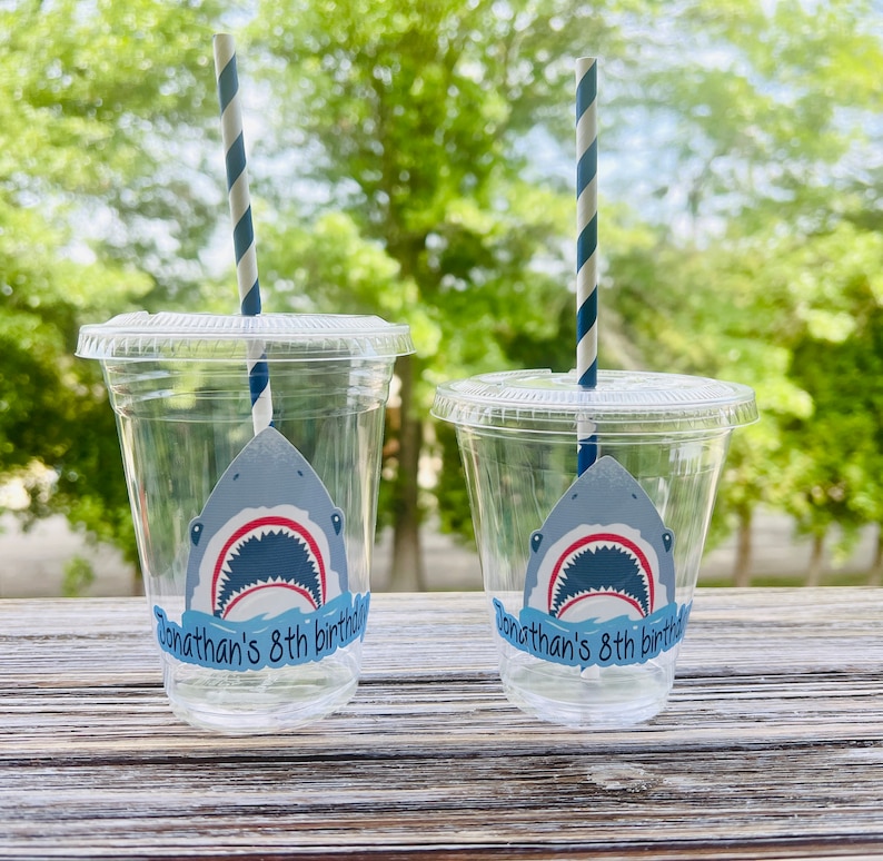 May include: Two clear plastic cups with blue and white striped straws. Each cup has a shark sticker with an open mouth and the text "Jonathan's 8th birthday" on a blue banner.