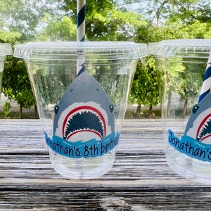 May include: Clear plastic cups with a shark graphic and the text "Jonathan's 8th birthday" on a blue banner. The cups are on a wooden surface.