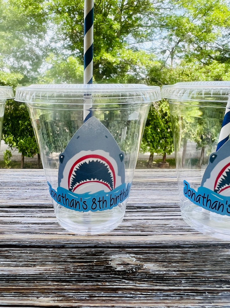 May include: Clear plastic cups with a shark graphic and the text "Jonathan's 8th birthday" on a blue banner. The cups are on a wooden surface.
