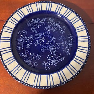 May include: A round ceramic plate with a navy blue center featuring white floral designs. The rim is cream-colored with vertical blue stripes and a dotted border. This decorative plate is suitable for serving food.