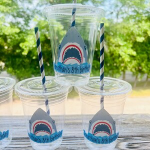 May include: Four clear plastic cups with blue and white striped straws. Each cup has a shark sticker with the text "Jonathan's 8th birthday".