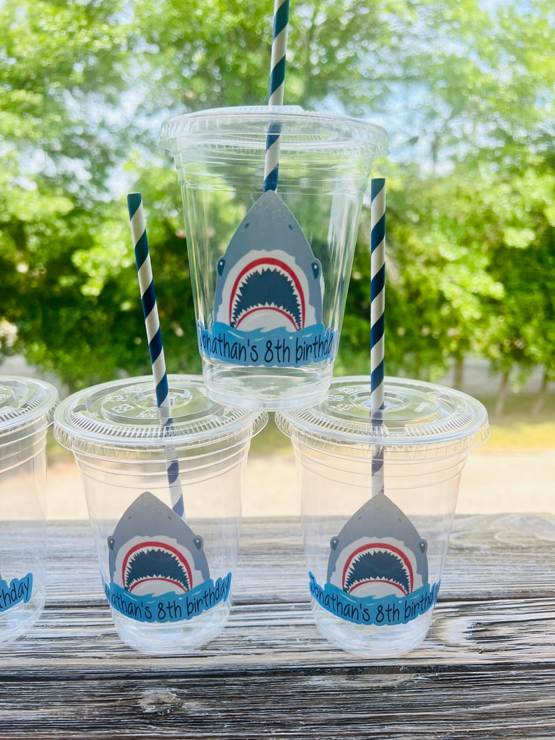 May include: Four clear plastic cups with blue and white striped straws. Each cup has a shark sticker with the text "Jonathan's 8th birthday".