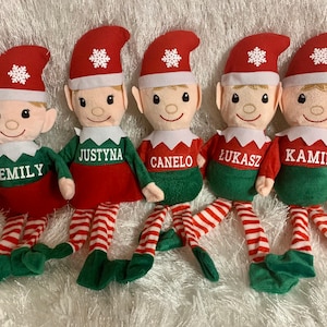 May include: Five plush Christmas elves with red and green outfits and white hats with snowflakes. Each elf has a different name embroidered on their shirt: Emily, Justyna, Canelo, Lukasz, and Kamil.