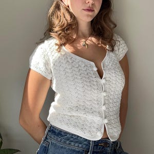 Może przedstawiać: Biały kardigan wykonany na szydełku z zapięciem na guziki i krótkimi rękawami. Kardigan ma delikatny, ażurowy wzór.