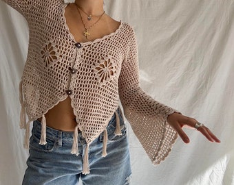 May include: A crocheted cardigan with a V-neck, long sleeves, and fringe detailing. The cardigan is a light beige color and has a button closure.