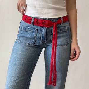 Può includere: Una cintura rossa all'uncinetto con un motivo floreale e lunghe nappe. La cintura è annodata intorno alla vita di una persona che indossa jeans blu e una canotta bianca. Un accessorio vivace.