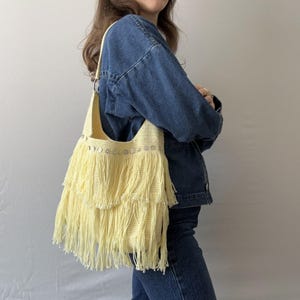 Puede incluir: Un bolso de hombro de crochet amarillo con detalles de flecos y adornos plateados.