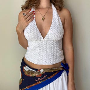 Puede incluir: Top blanco con cuello halter y escote en V profundo. La parte superior está hecha de una tela texturizada de tejido abierto. Se ve un collar dorado con un colgante. Una bufanda estampada en tonos azules, rojos y dorados está atada a la cintura.