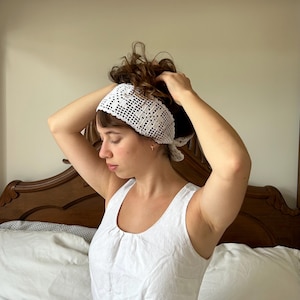 Puede incluir: Una diadema blanca de ganchillo con un diseño calado. La diadema está atada en la parte posterior. La persona de la imagen lleva una camiseta sin mangas blanca. El fondo es un dormitorio.
