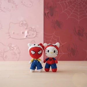 Peut inclure: Deux poupées au crochet représentant des visages de chat blancs avec moustaches et nœuds, vêtues de tenues de super-héros rouges et bleues. Une poupée porte un masque rouge avec des yeux blancs, l'autre une capuche rouge à motif toile d'araignée.
