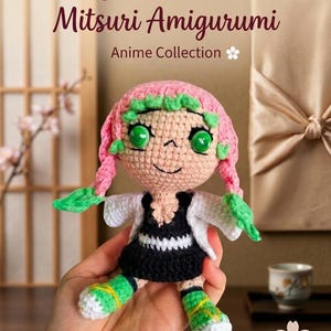 May include: Handmade Mitsuri Amigurumi doll from the Anime Collection. The crochet doll features pink hair with green accents, large green eyes, and a black and white dress. The doll is held in a hand.