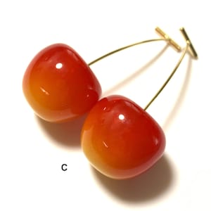 K&ouml;nnte beinhalten: Ein Paar Kirsch-Ohrringe mit goldfarbenem Draht und einem T-f&ouml;rmigen Verschluss. Die Kirschen sind rot und orange mit einem Farbverlauf.