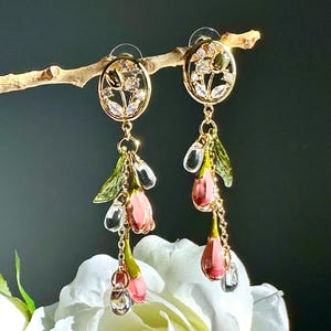 May include: Gold-toned floral earrings with dangling pink flower bud charms and clear crystal accents. The earrings feature an oval-shaped top with a floral design and are suspended from a delicate chain. The earrings are displayed on a branch.