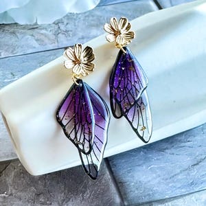 May include: A pair of dangle earrings featuring a gold flower stud and purple, wing-shaped pendants. The pendants have a black outline and a gradient of purple and clear colors, with gold flecks. The earrings are displayed on a white surface.