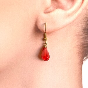 Pode incluir: Um &uacute;nico brinco dourado com uma conta vermelha transl&uacute;cida em forma de gota. O brinco est&aacute; pendurado em uma orelha, mostrando o design delicado e a cor da joia. O gancho e a conta do brinco s&atilde;o claramente vis&iacute;veis.