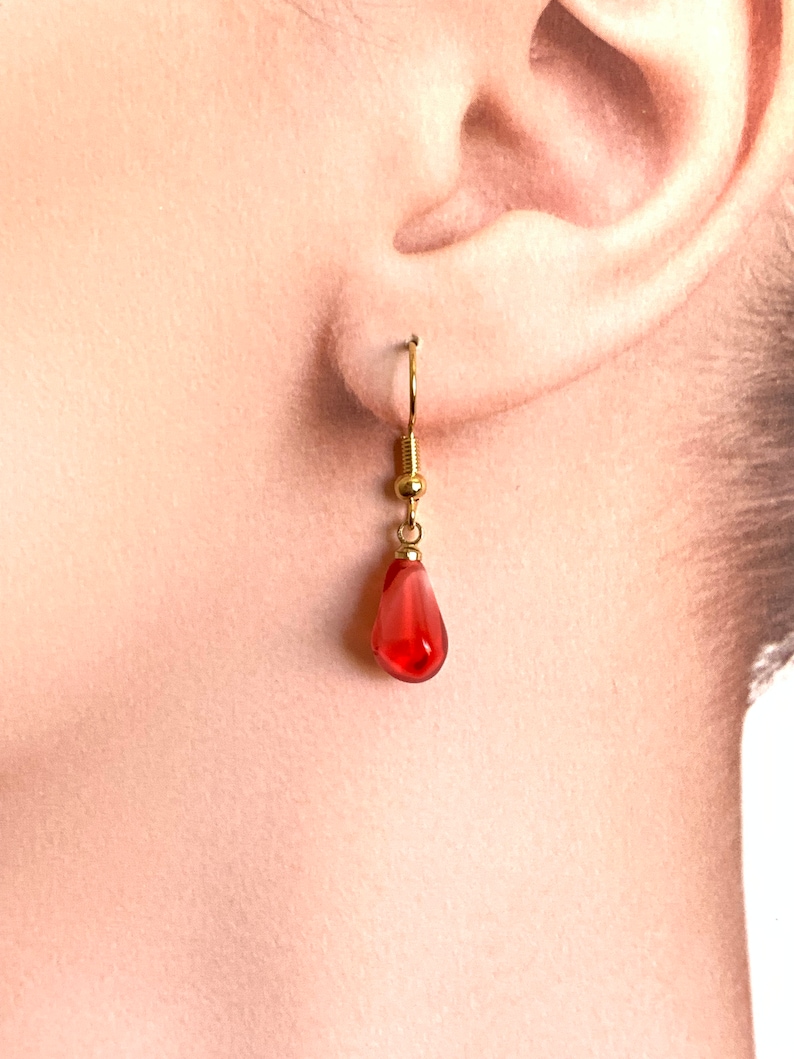 Pode incluir: Um &uacute;nico brinco dourado com uma conta vermelha transl&uacute;cida em forma de gota. O brinco est&aacute; pendurado em uma orelha, mostrando o design delicado e a cor da joia. O gancho e a conta do brinco s&atilde;o claramente vis&iacute;veis.