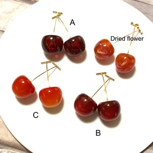 K&ouml;nnte beinhalten: Vier Paar Kirsch-Ohrringe, jedes Paar in einer anderen Farbe und einem anderen Stil. Die Ohrringe sind aus Harz und haben goldfarbene Dr&auml;hte. Die Ohrringe sind mit A, B, C und Trockenblume beschriftet.