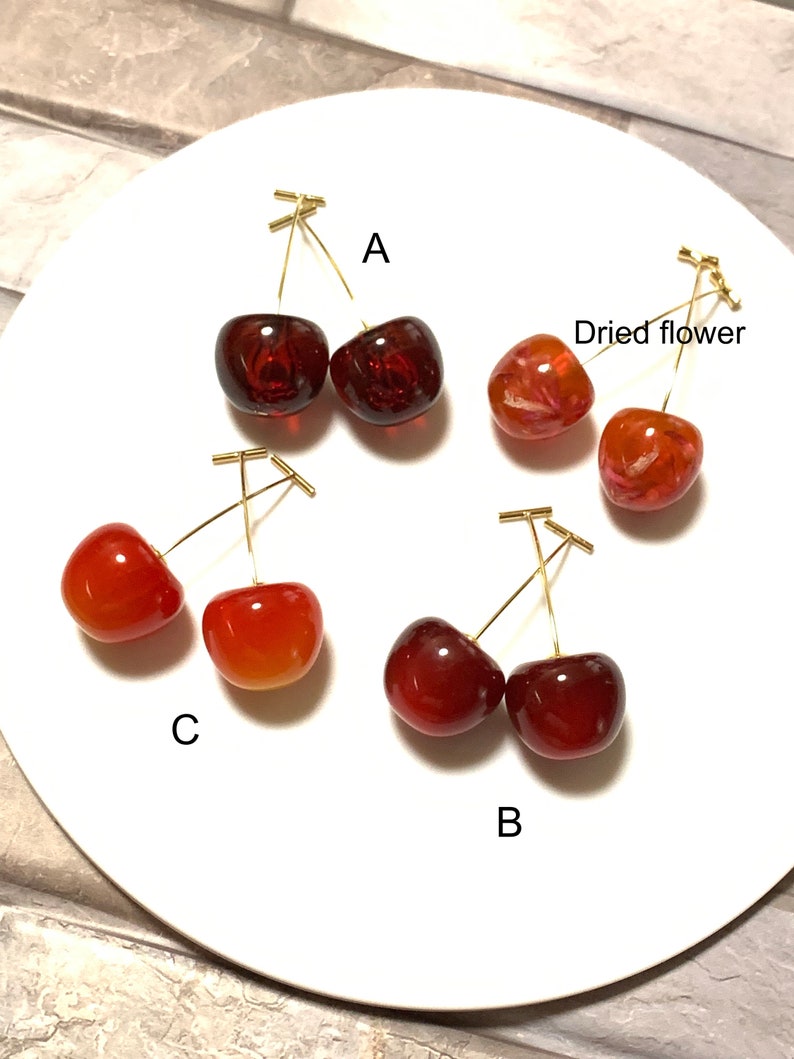 K&ouml;nnte beinhalten: Vier Paar Kirsch-Ohrringe, jedes Paar in einer anderen Farbe und einem anderen Stil. Die Ohrringe sind aus Harz und haben goldfarbene Dr&auml;hte. Die Ohrringe sind mit A, B, C und Trockenblume beschriftet.