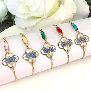 May include: Five gold-tone bracelets featuring a four-leaf clover charm filled with clear resin and pressed blue forget-me-not flowers. Each bracelet includes a single oval-shaped crystal in pink, yellow, green, red, or blue attached to the chain.