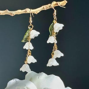 May include: Dangling earrings featuring white flower charms and green leaf accents. The earrings are suspended from gold-colored hooks and chains. The floral design is delicate and detailed, with a natural aesthetic. The earrings are displayed against a dark background.
