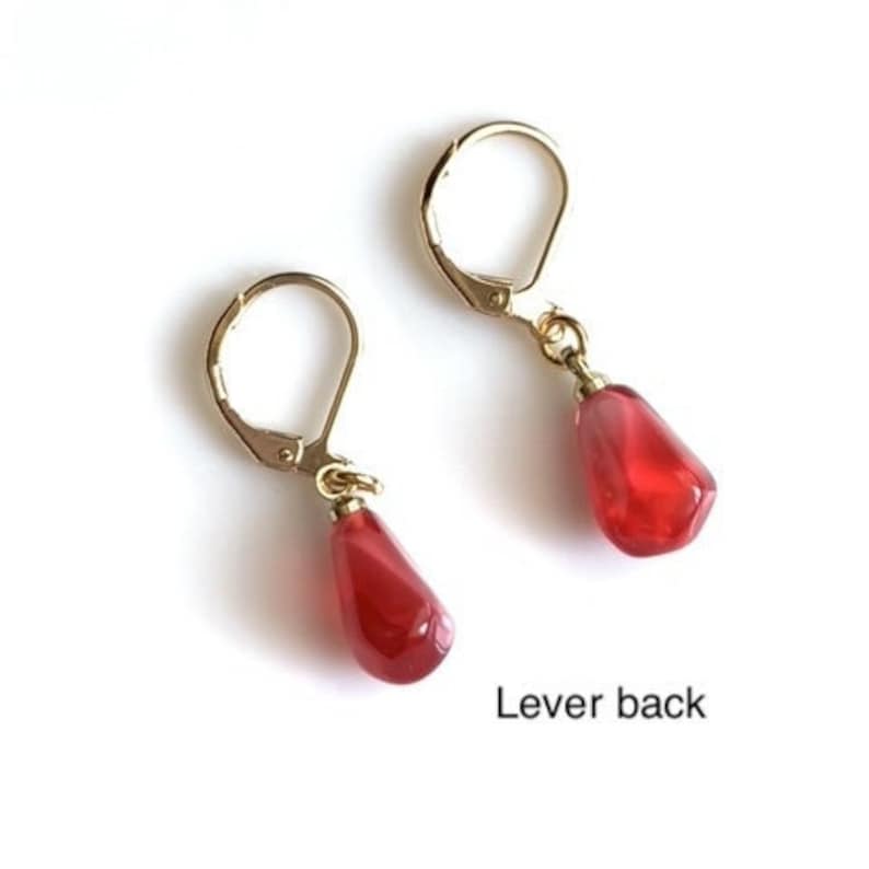 Pode incluir: Um par de brincos dourados com fecho de alavanca, cada um com uma conta vermelha transl&uacute;cida em forma de gota. Os brincos s&atilde;o exibidos em um fundo branco. O texto "Lever back" &eacute; vis&iacute;vel.