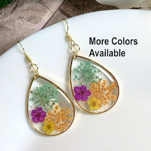 May include: Teardrop-shaped earrings featuring clear resin frames with gold-tone metal edges. Each earring contains pressed dried flowers in shades of purple, yellow, orange, and light green. The text More Colors Available is printed on the right.
