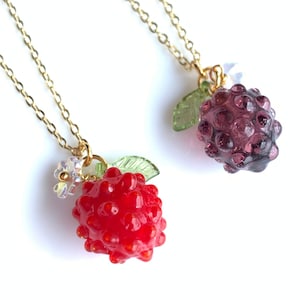 May include: Two gold necklaces with glass berry charms. One necklace has a red berry charm with a green leaf and a white flower. The other necklace has a purple berry charm with a green leaf.