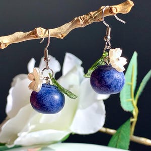 May include: A pair of dangle earrings featuring dark blue, blueberry-shaped beads. Each earring has a small white flower and a green leaf accent. The earrings hang from silver-colored fish hook ear wires, displayed on a small branch against a dark background.
